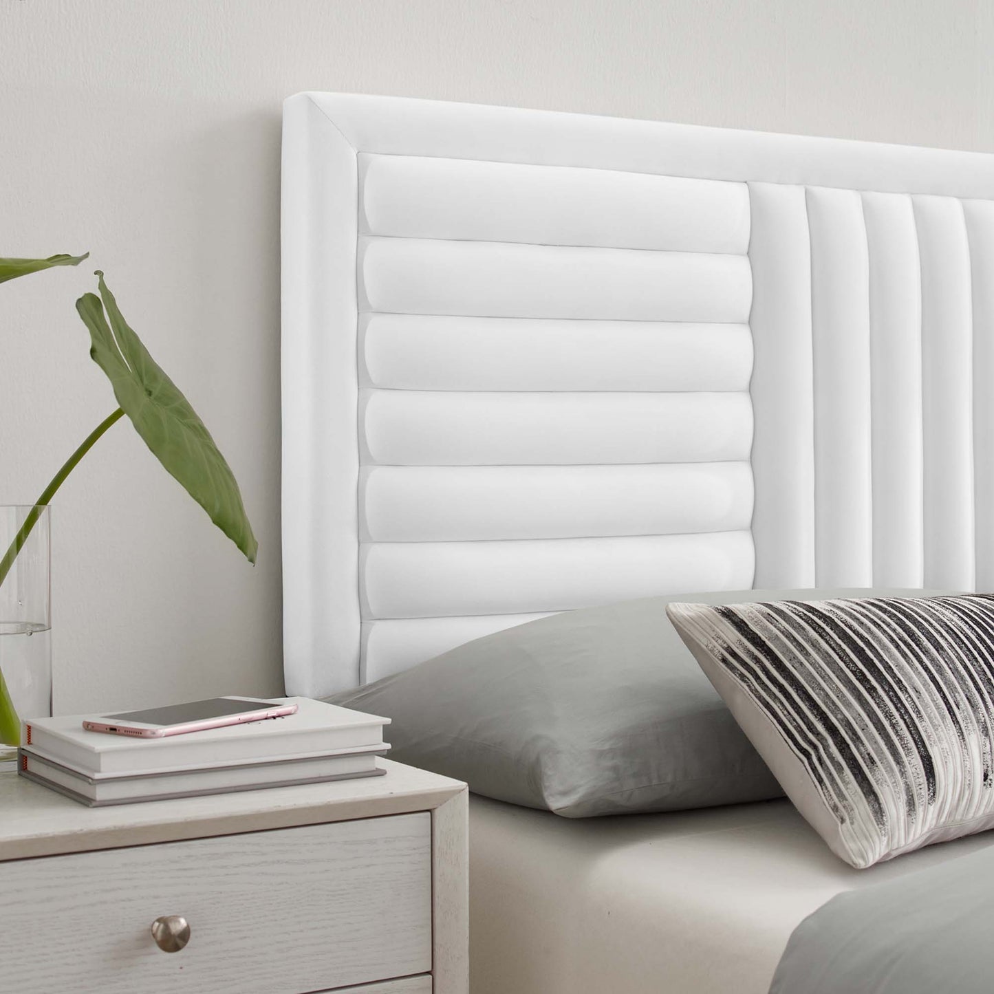 White upholstered headboard with vertical channel tufting, gray bedding, striped pillow, nightstand with books and plant