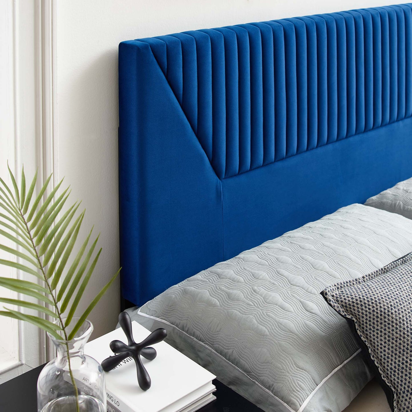 Navy blue velvet headboard with vertical channel tufting, gray quilted bedding, white wall, glass vase with a plant.