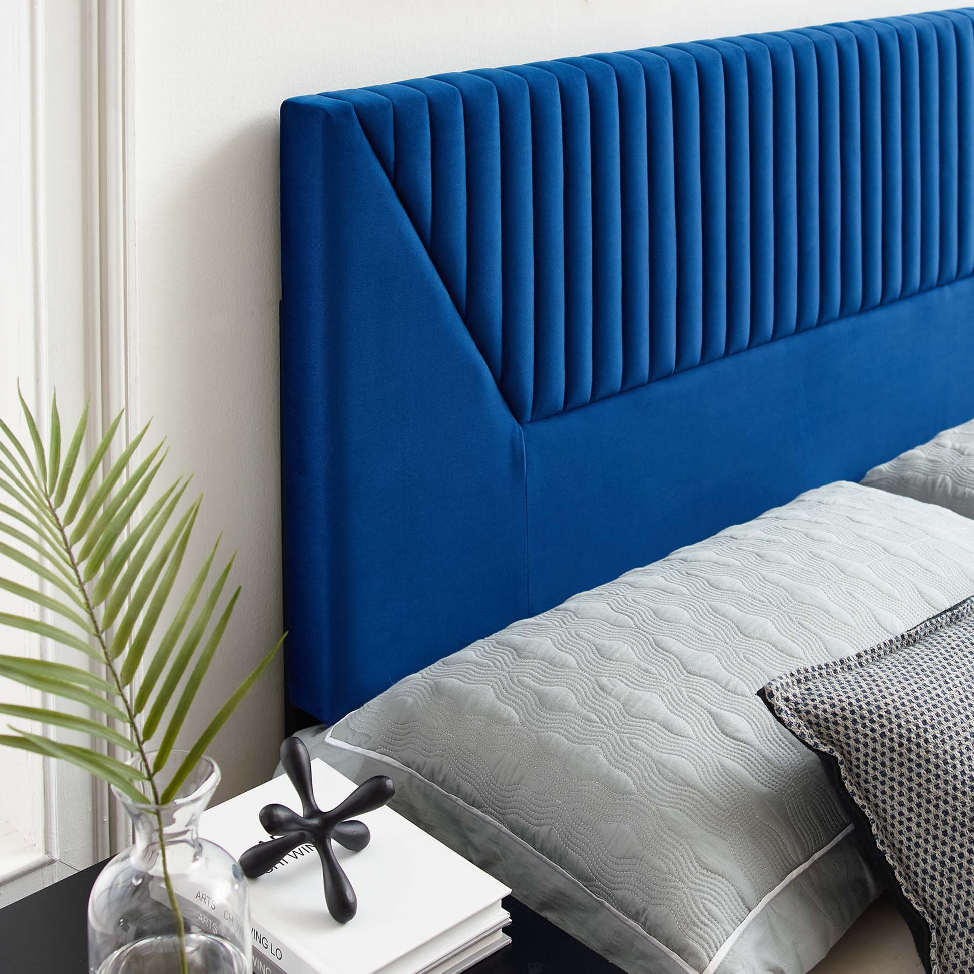 Navy blue velvet headboard with vertical channel tufting, gray quilted bedding, white wall, glass vase with a plant.
