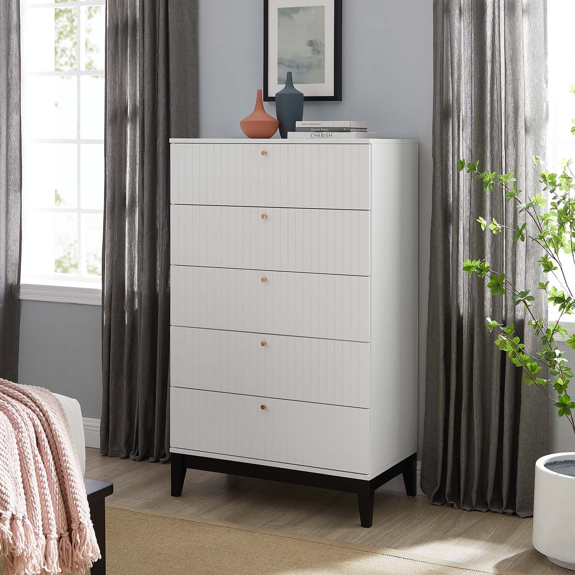 White five-drawer dresser with vertical grooves, wooden knobs, black tapered legs; decor on top in a modern bedroom.