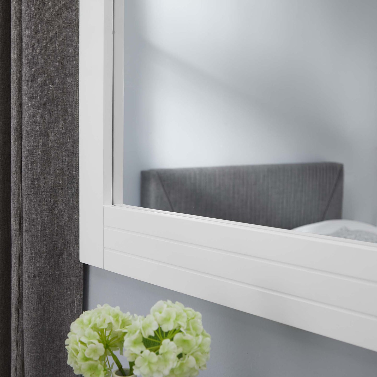 White framed wall mirror with clean lines; reflects gray headboard, dark curtain, and pale green hydrangea in foreground.