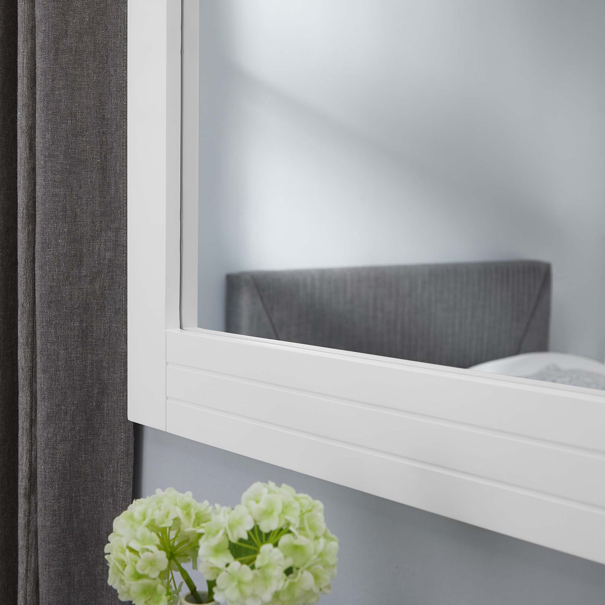 White framed wall mirror with clean lines; reflects gray headboard, dark curtain, and pale green hydrangea in foreground.