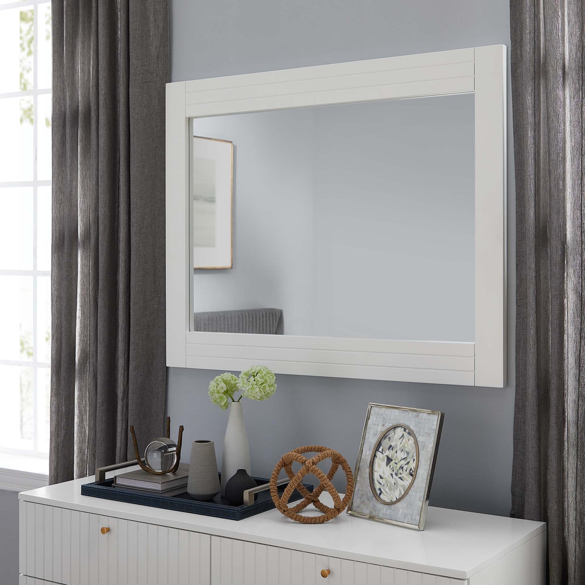 White-framed rectangular mirror above a white dresser with gold knobs; vase of green flowers, wicker knot sculpture, and framed art.