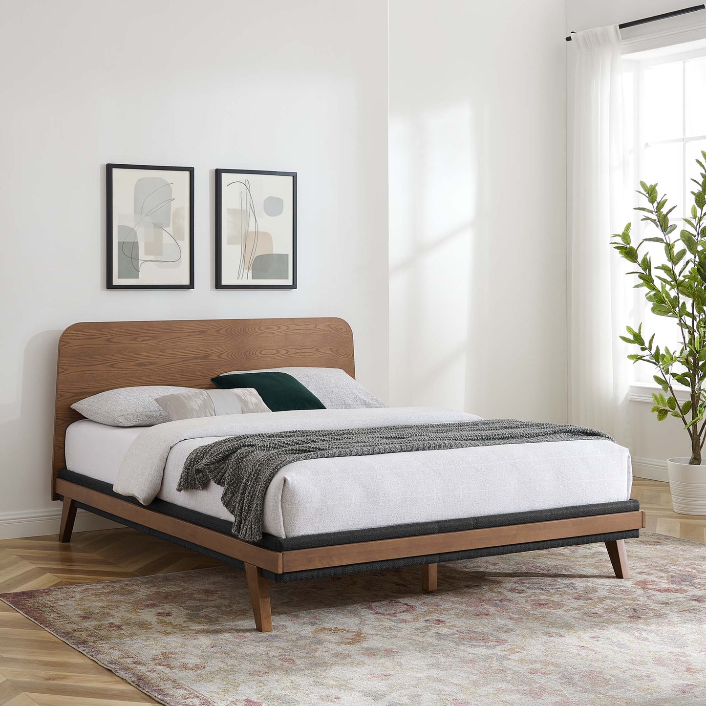 Mid-century walnut bed with curved headboard, white linens, gray throw, green pillow, abstract framed art, and potted plant.