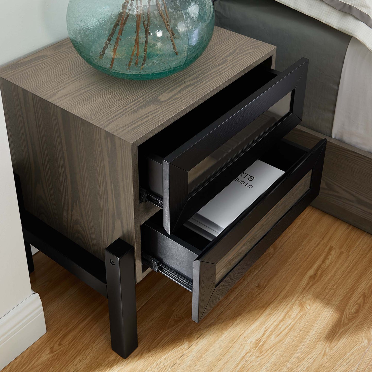 Oak wood nightstand with black metal frame & two open drawers; glass vase on top.