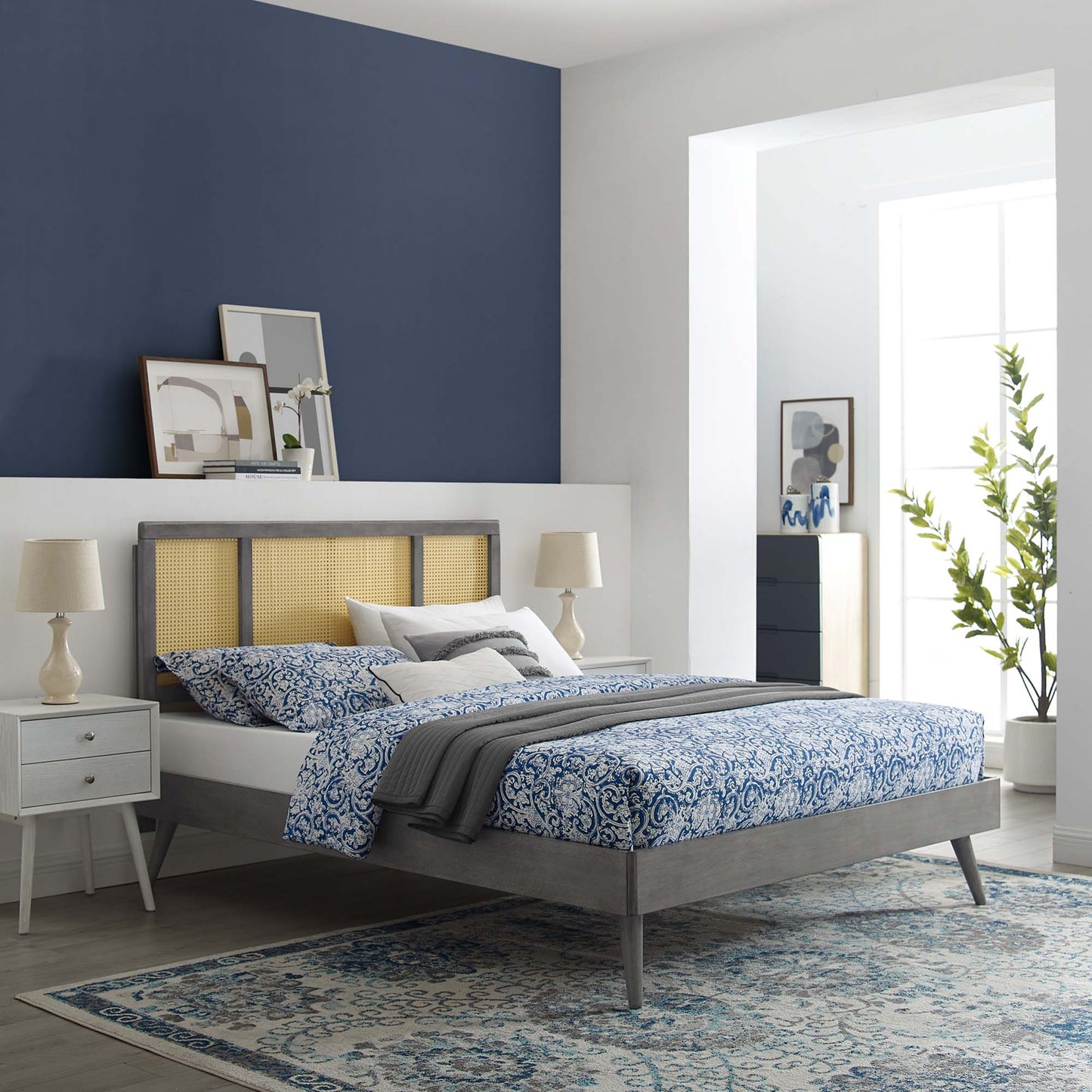 Grey wood bed frame with cane headboard & blue damask bedding, gray throw, white pillows & beige lamps, blue accent wall.