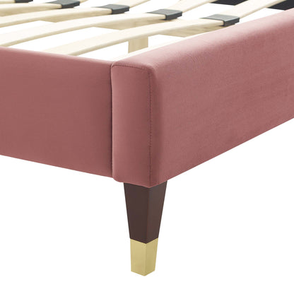 Dusty-rose upholstered bed frame & a dark wood leg capped in brass; slatted base visible.