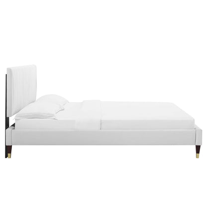 White velvet upholstered full platform bed with a tall padded headboard, white linens and gold-tipped legs.