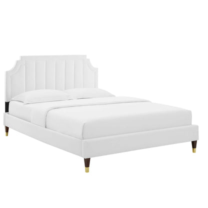 White upholstered platform bed with mattress, vertical channel tufted headboard, and tapered wooden legs with brass caps.