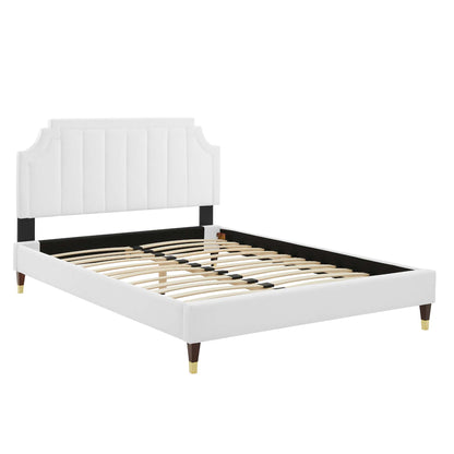 White upholstered platform bed with tall vertically tufted headboard, black rails, wooden slats, & gold-tipped legs.