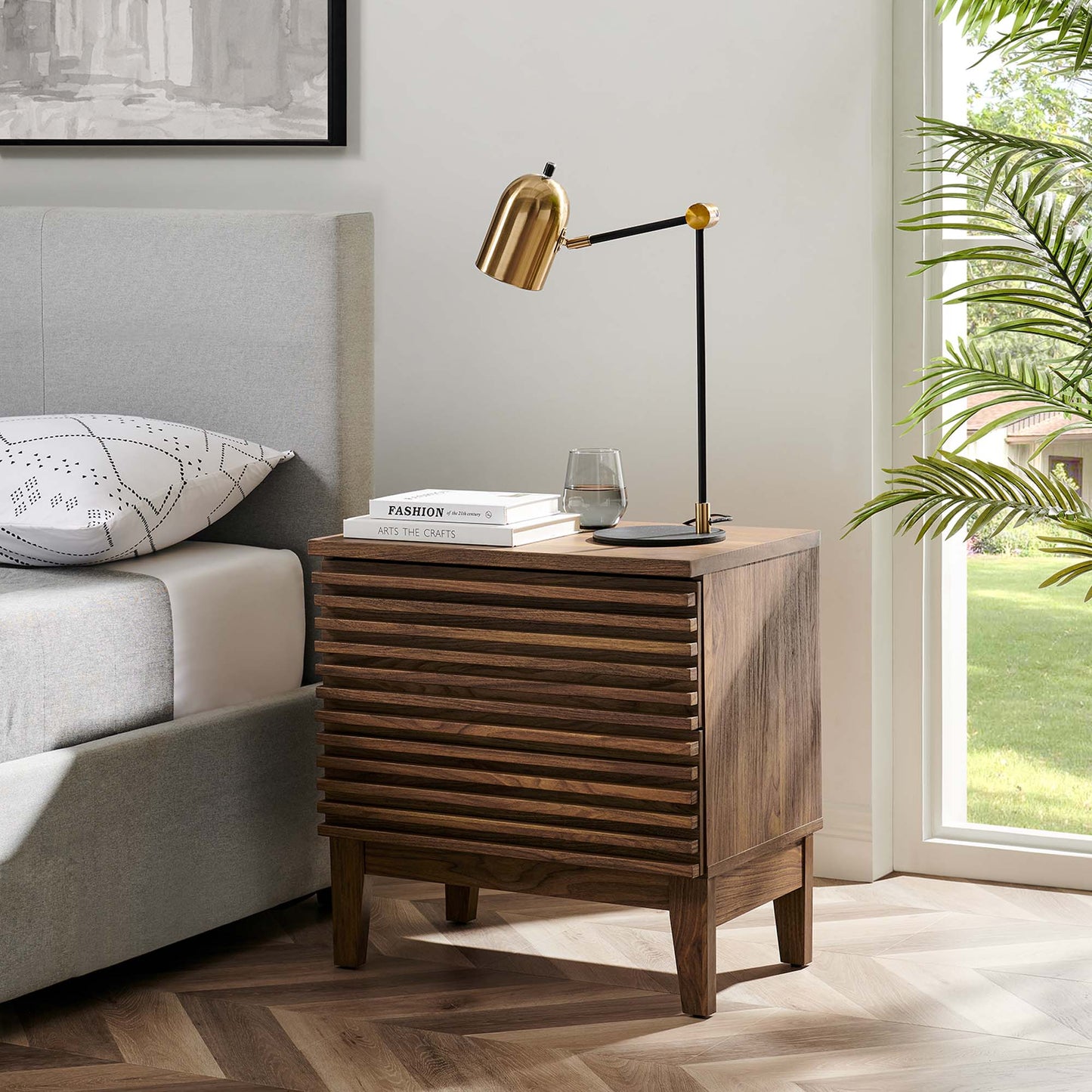 Walnut two-drawer mid-century nightstand with slatted front, brass adjustable lamp, books and glass atop beside a gray upholstered bed.