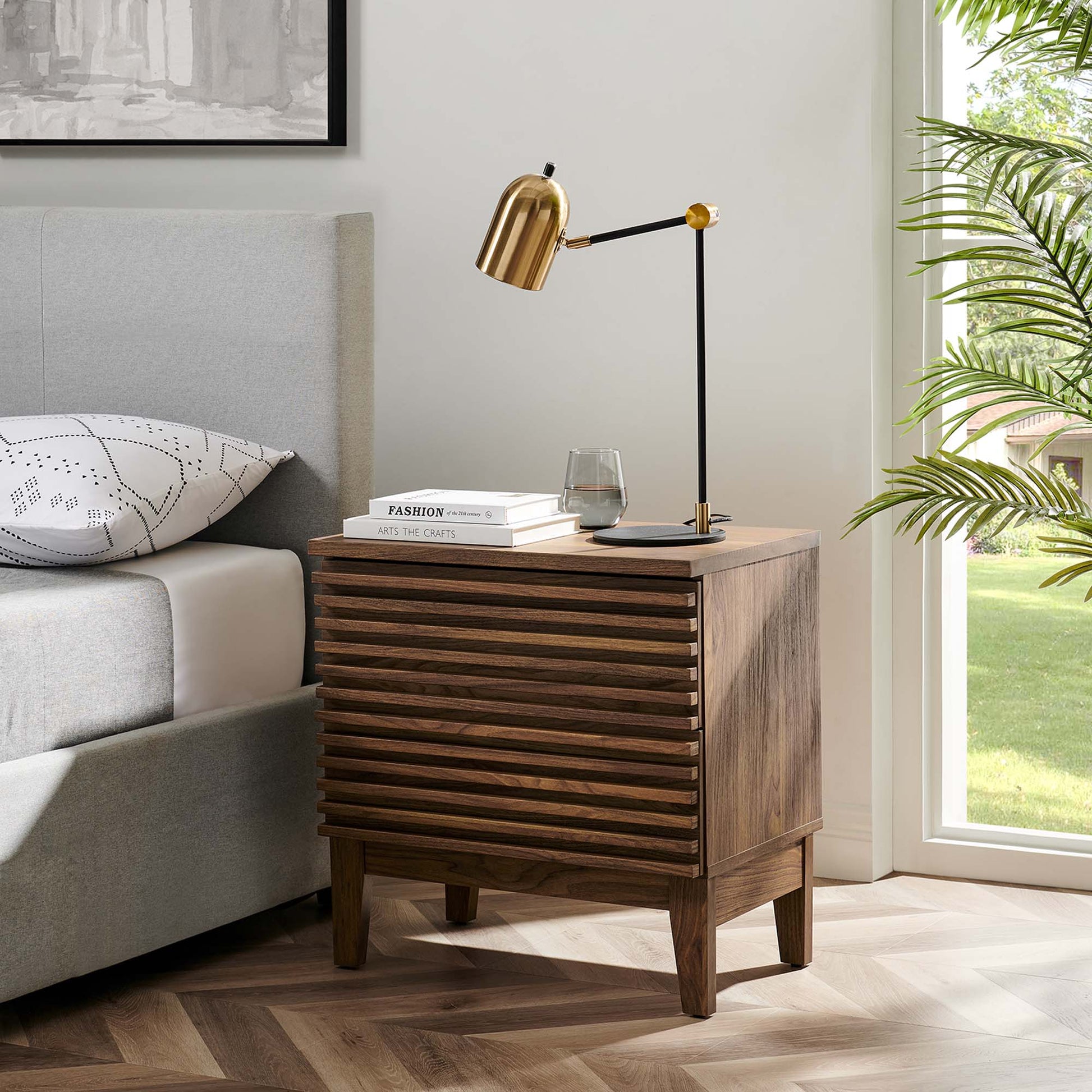 Walnut two-drawer mid-century nightstand with slatted front, brass adjustable lamp, books and glass atop beside a gray upholstered bed.