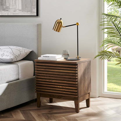 Walnut two-drawer mid-century nightstand with slatted front, brass adjustable lamp, books and glass atop beside a gray upholstered bed.