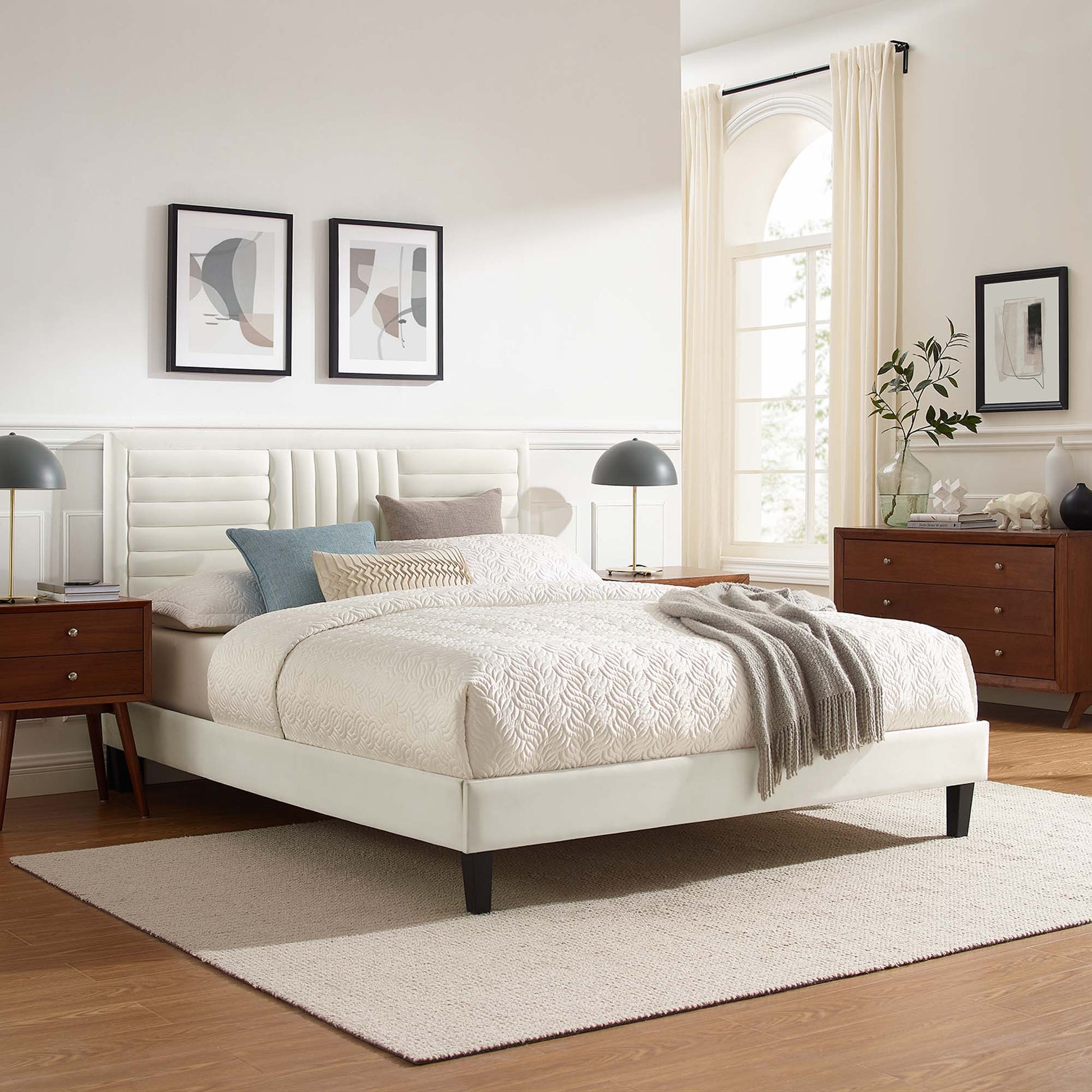White upholstered platform bed with tufted headboard, cream bedding, blue accent pillows, gray throw, dark wood nightstands & dresser.