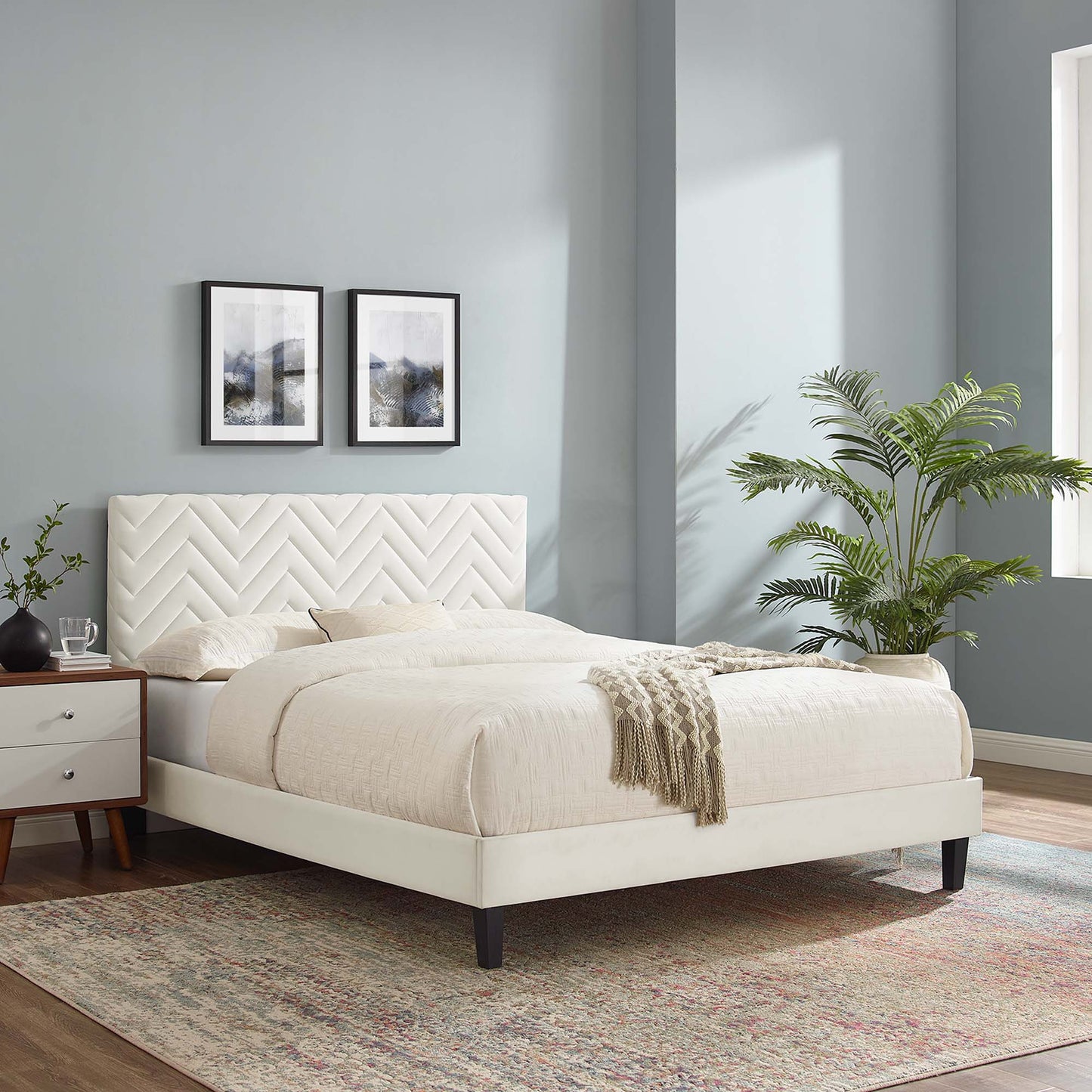 White upholstered platform bed with chevron tufted headboard, beige bedding, white nightstands, potted palm, blue-gray walls.
