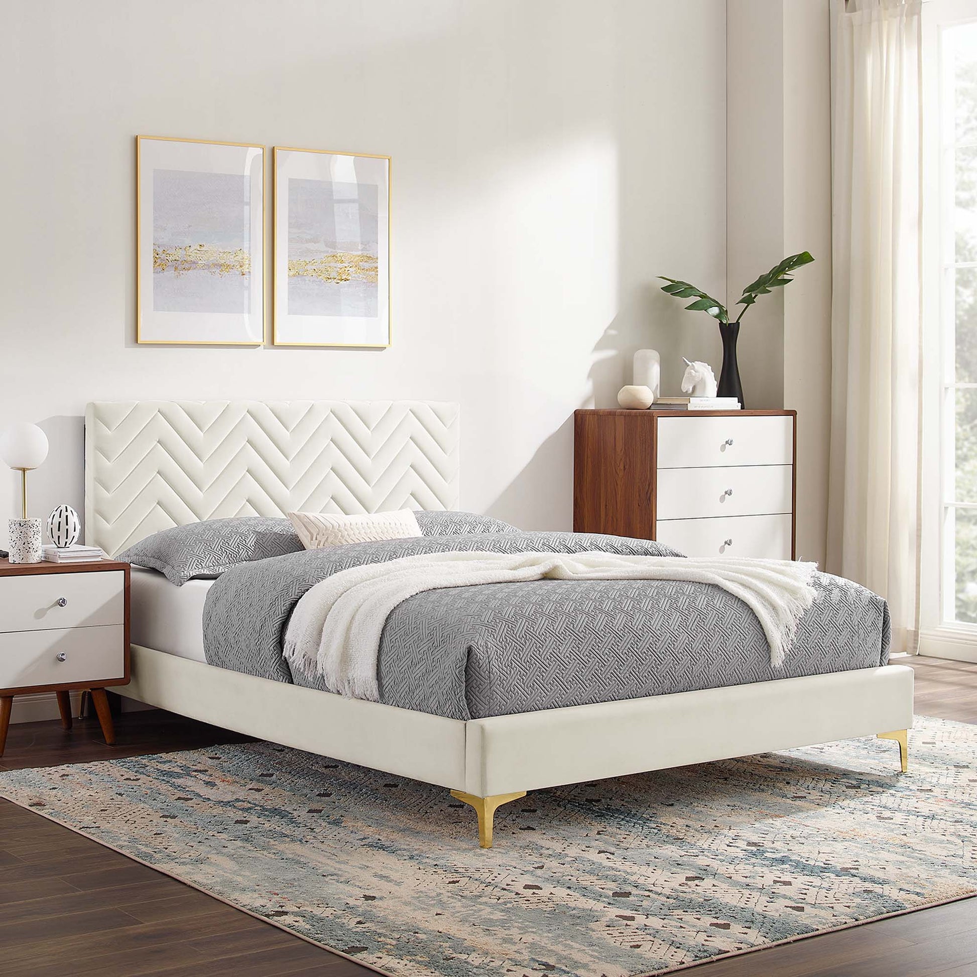 Cream upholstered platform bed with chevron-tufted headboard, gray bedding, gold legs; wood nightstands and dresser.