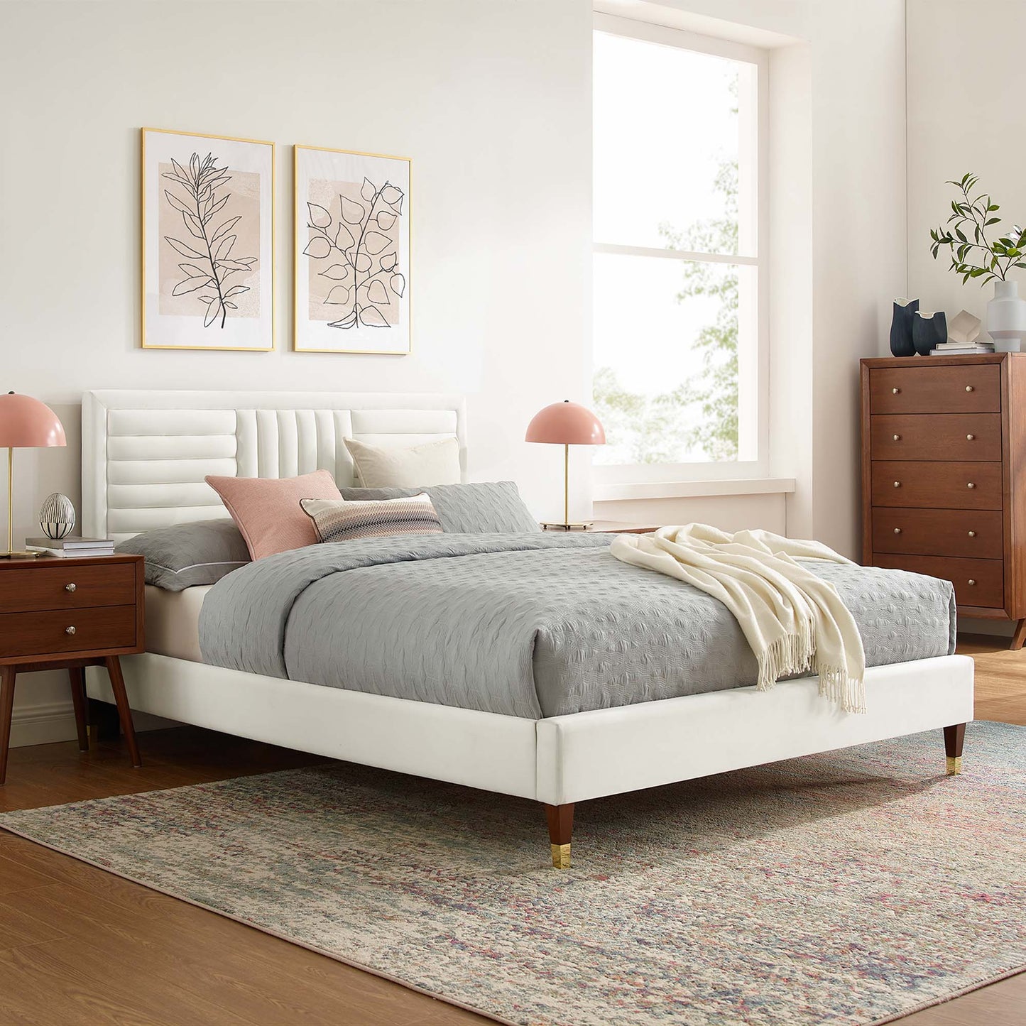 White upholstered platform bed with gray bedding, pink pillows, wood nightstands, dresser, pastel rug, botanical art.