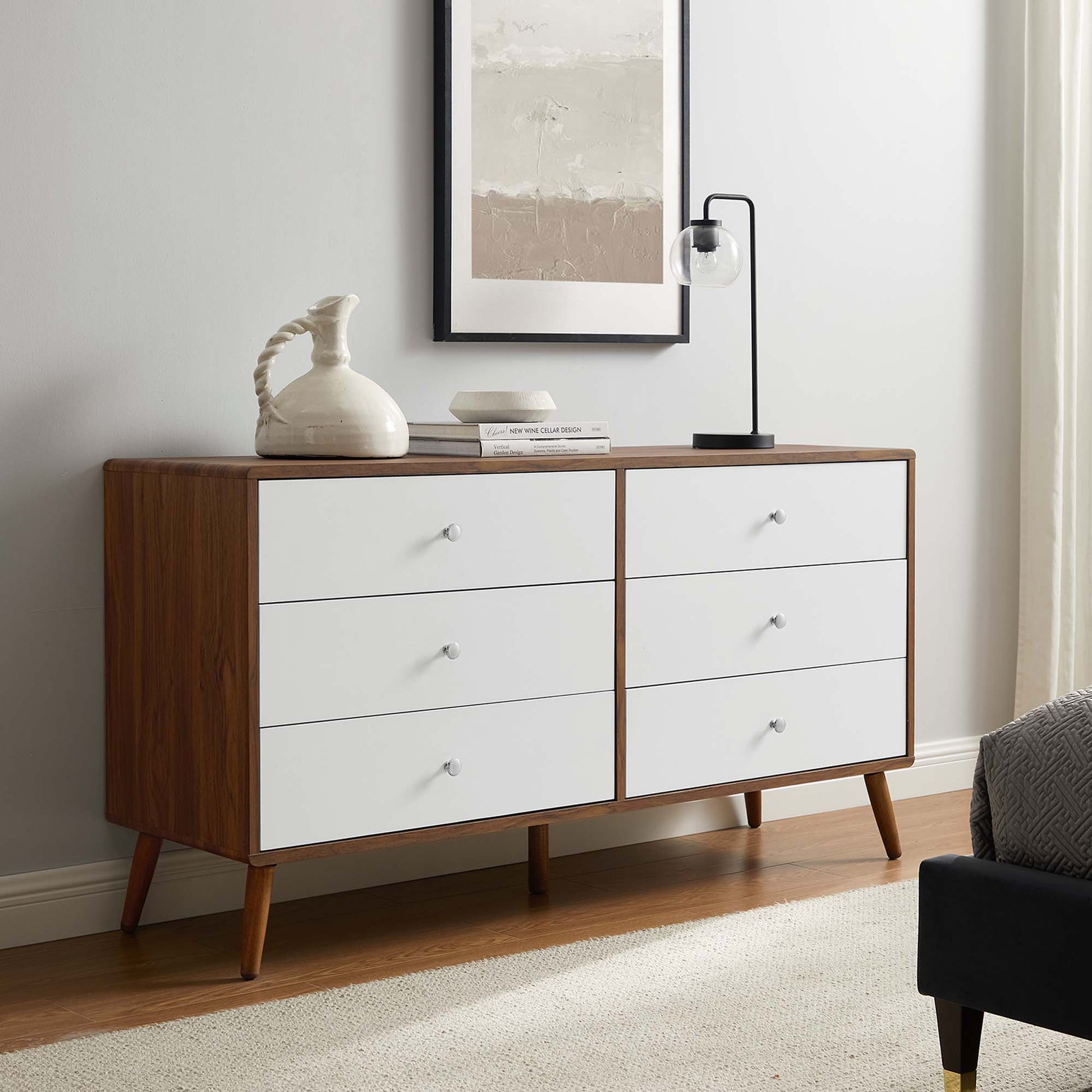 Mid-century white dresser with six drawers, ceramic pitcher, books and a black glass lamp, framed art.