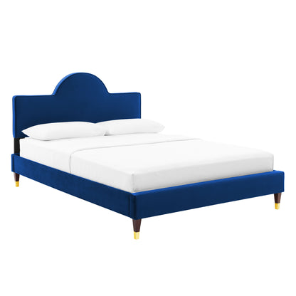 Navy blue velvet bed with curved arched headboard, low platform frame, tapered wooden legs with gold caps, white bedding.