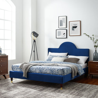 Navy velvet bed with curved headboard, blue-white damask bedding, gray throw, gold-tipped legs; paired with wooden nightstands.