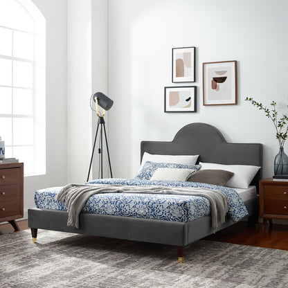 Charcoal upholstered bed with arched headboard, blue damask bedding, knit throw; wood nightstands, tripod lamp, abstract wall art.