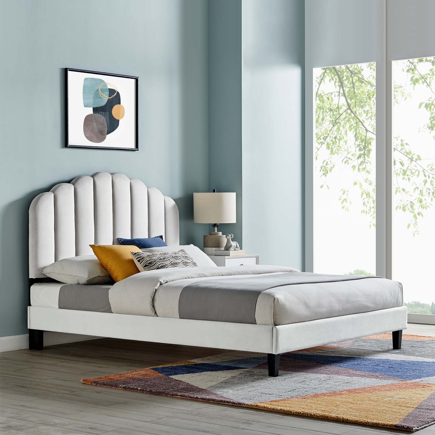 Modern upholstered bed with scalloped light-gray headboard, gray bedding, yellow and blue pillows, abstract art, colorful rug