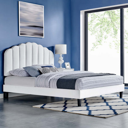 White bed with scalloped headboard, dark legs, grey bedding, blue walls, nightstand, lamp, and blue-and-white rug.