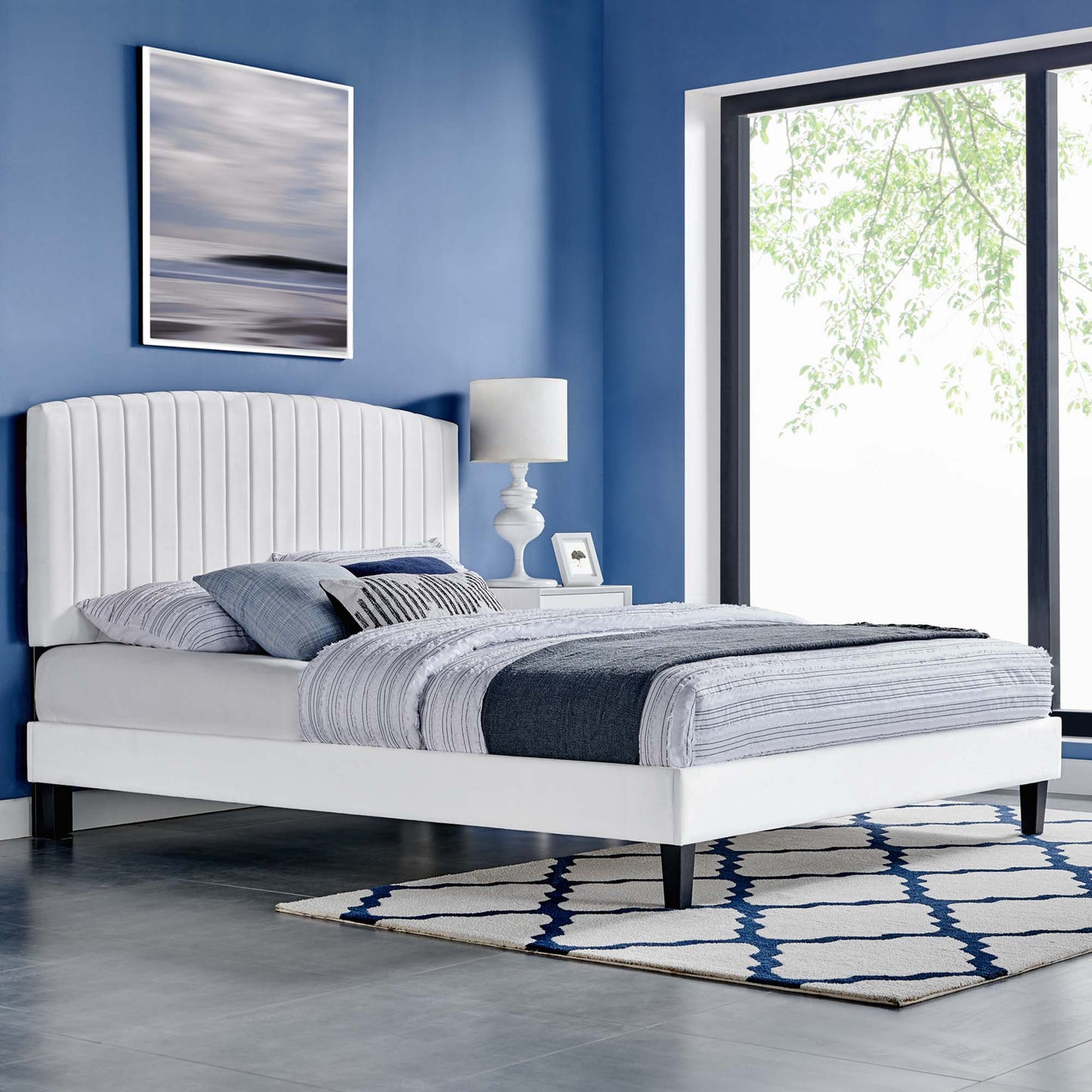 White platform bed with tall upholstered headboard in blue room, striped gray-blue bedding, modern lamp, rug, large window.