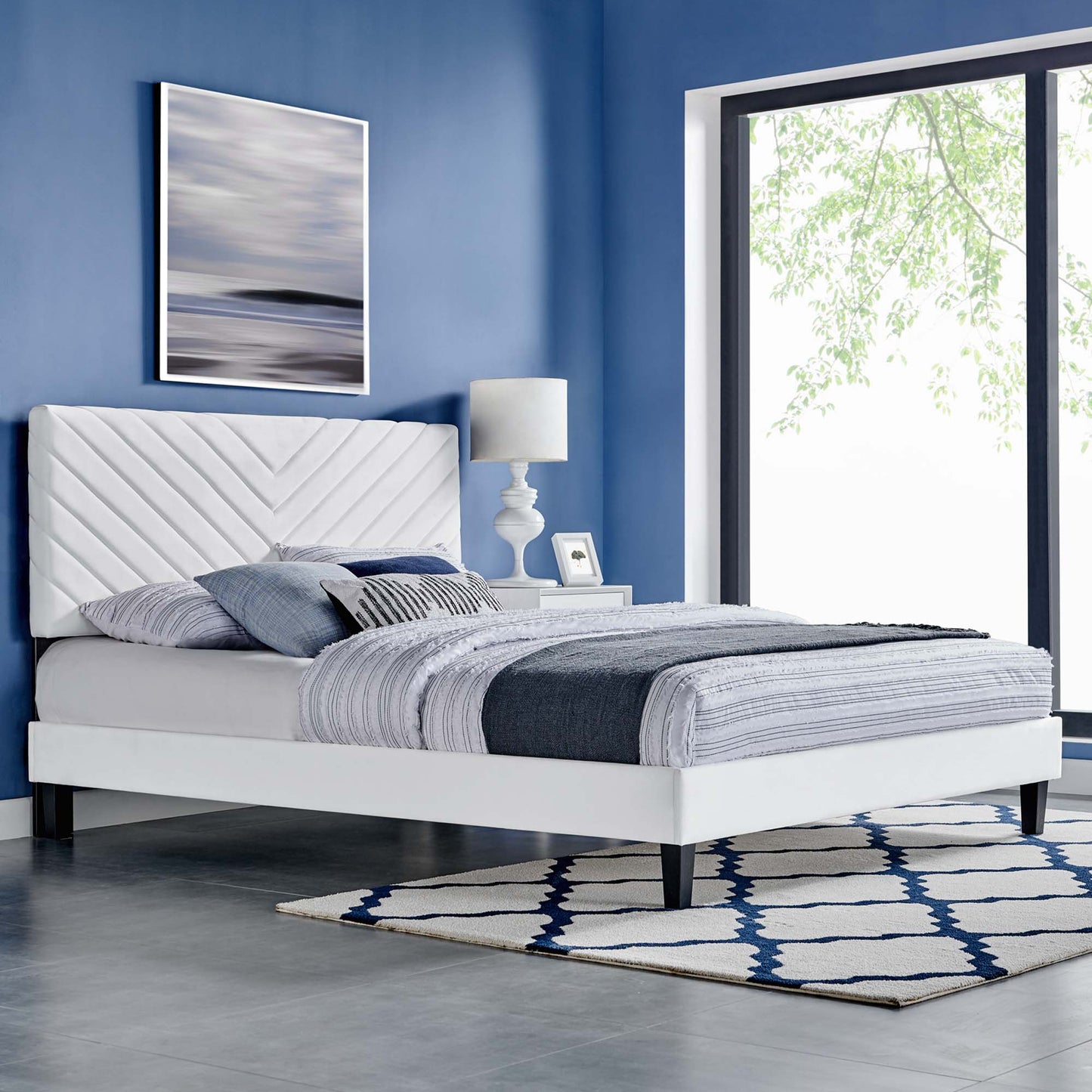 White platform bed with diagonally upholstered headboard, gray-blue linens, blue walls, nightstand lamp, and blue chevron rug.