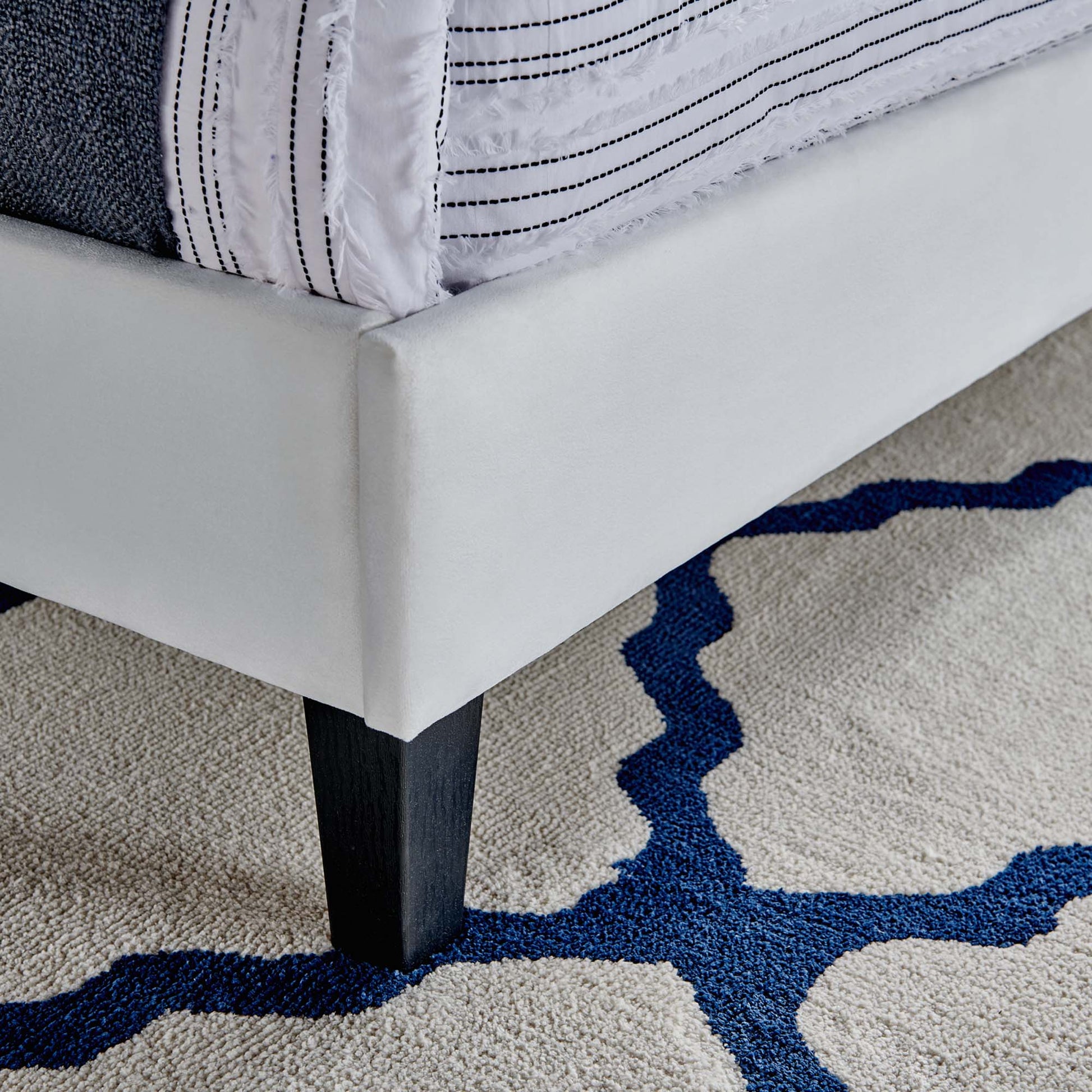 White upholstered bed frame with dark wood legs, striped white bedding, on a beige rug with a blue abstract pattern.