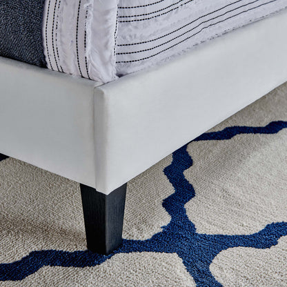 White upholstered bed frame with dark wood legs, striped white bedding, on a beige rug with a blue abstract pattern.