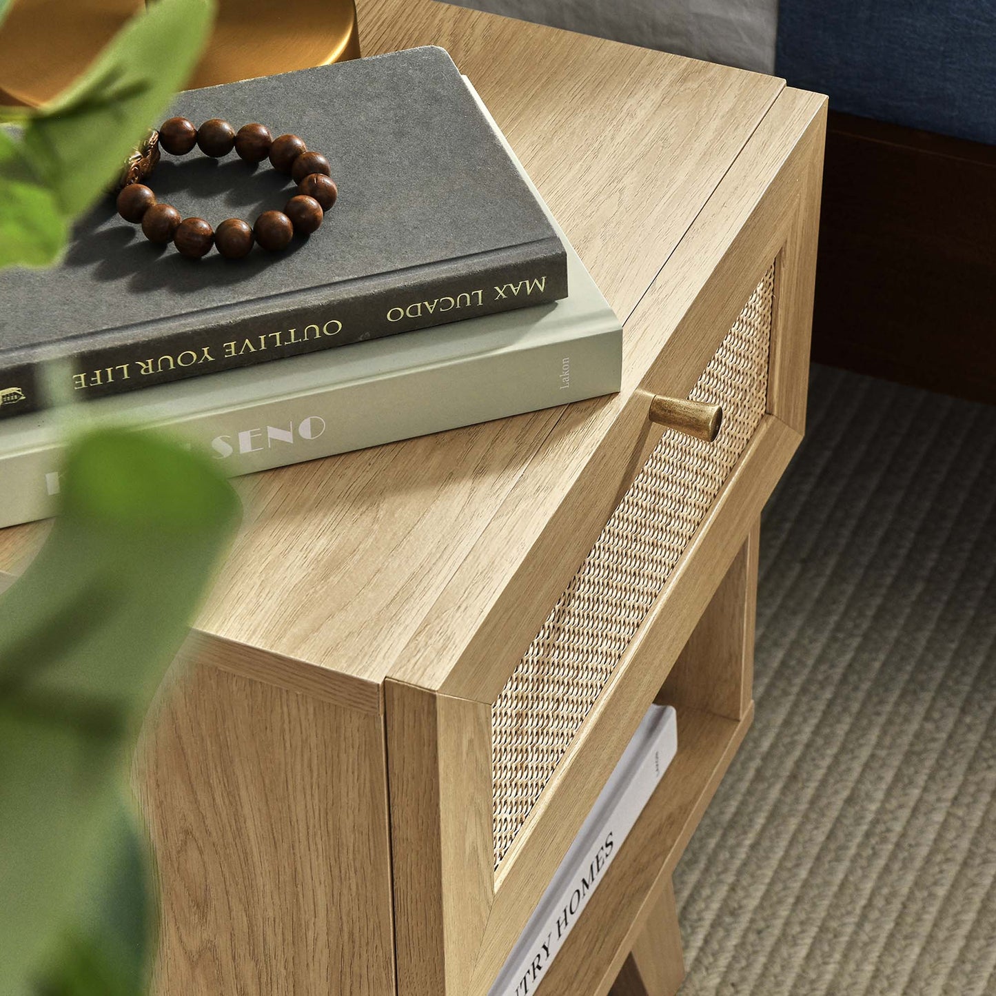 Natural oak nightstand with a woven cane drawer front and brass knob, 1-drawer, topped with books and a wooden bead bracelet.