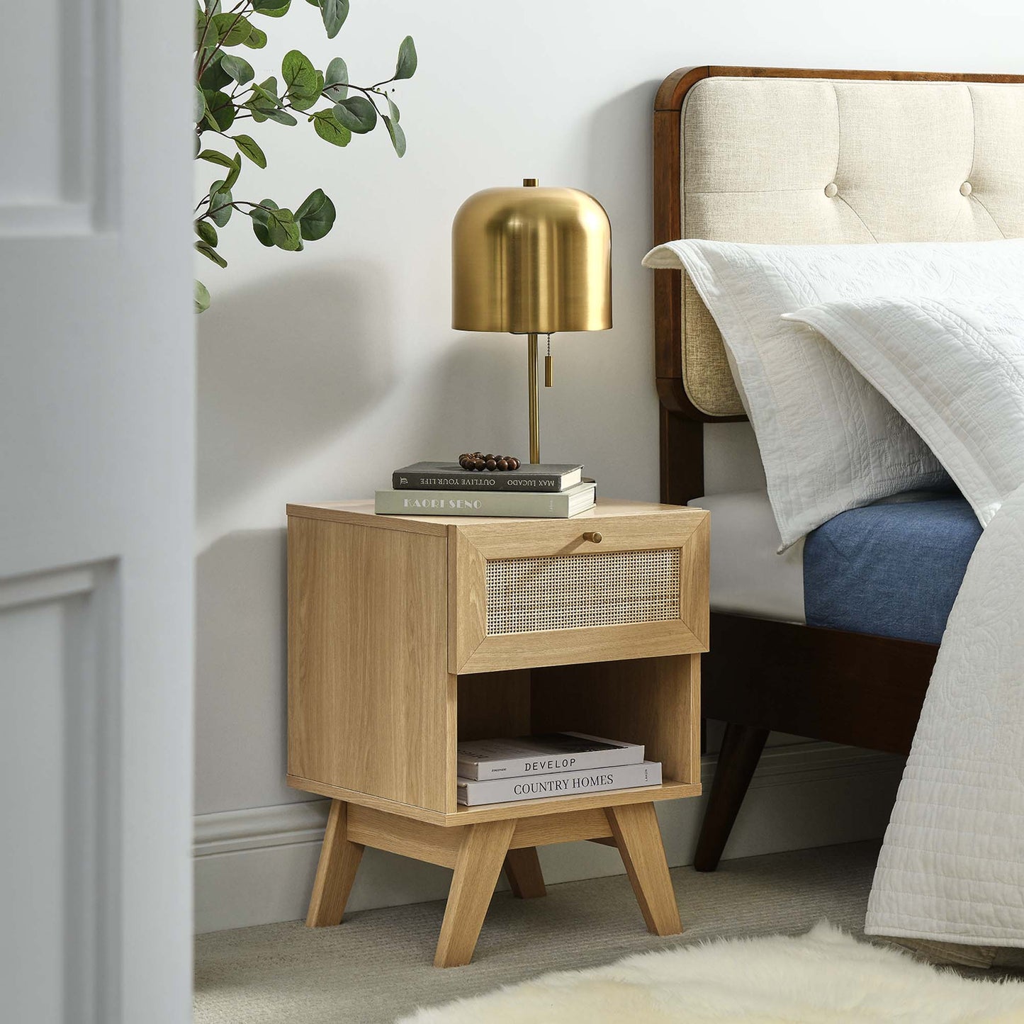 Oak nightstand with woven-front drawer, open lower shelf and angled legs; gold lamp and books in a cozy bedroom.