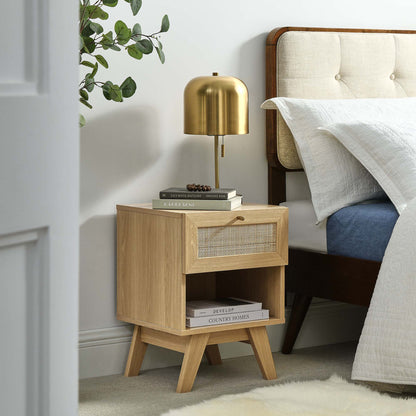 Oak nightstand with woven-front drawer, open lower shelf and angled legs; gold lamp and books in a cozy bedroom.