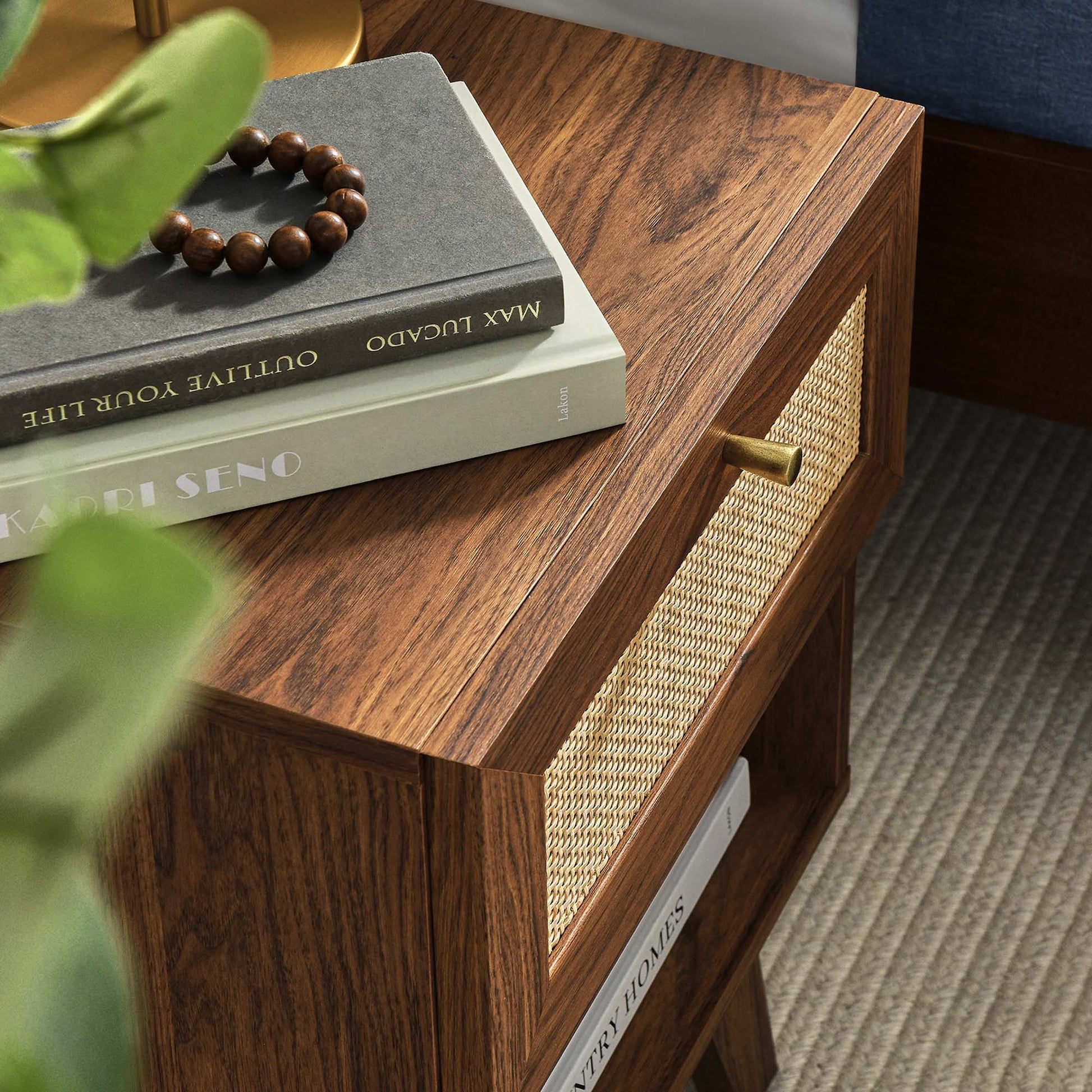 Walnut wood nightstand with a woven cane drawer front and brass knob; books and a beaded bracelet on top.