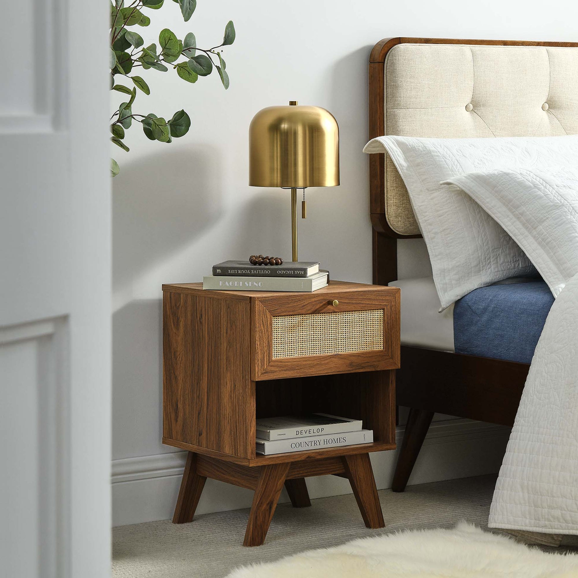 Walnut mid-century nightstand with woven rattan drawer, tapered legs, gold dome lamp on top, beside a bed.