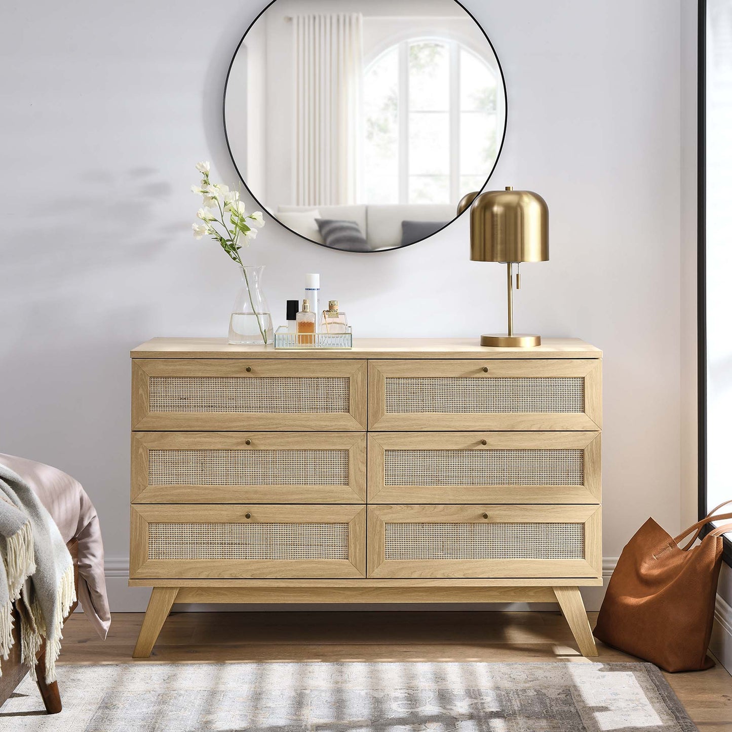 Oak dresser with six cane-front drawers & mid-century legs; brass lamp, vase & bottles on top under round mirror.