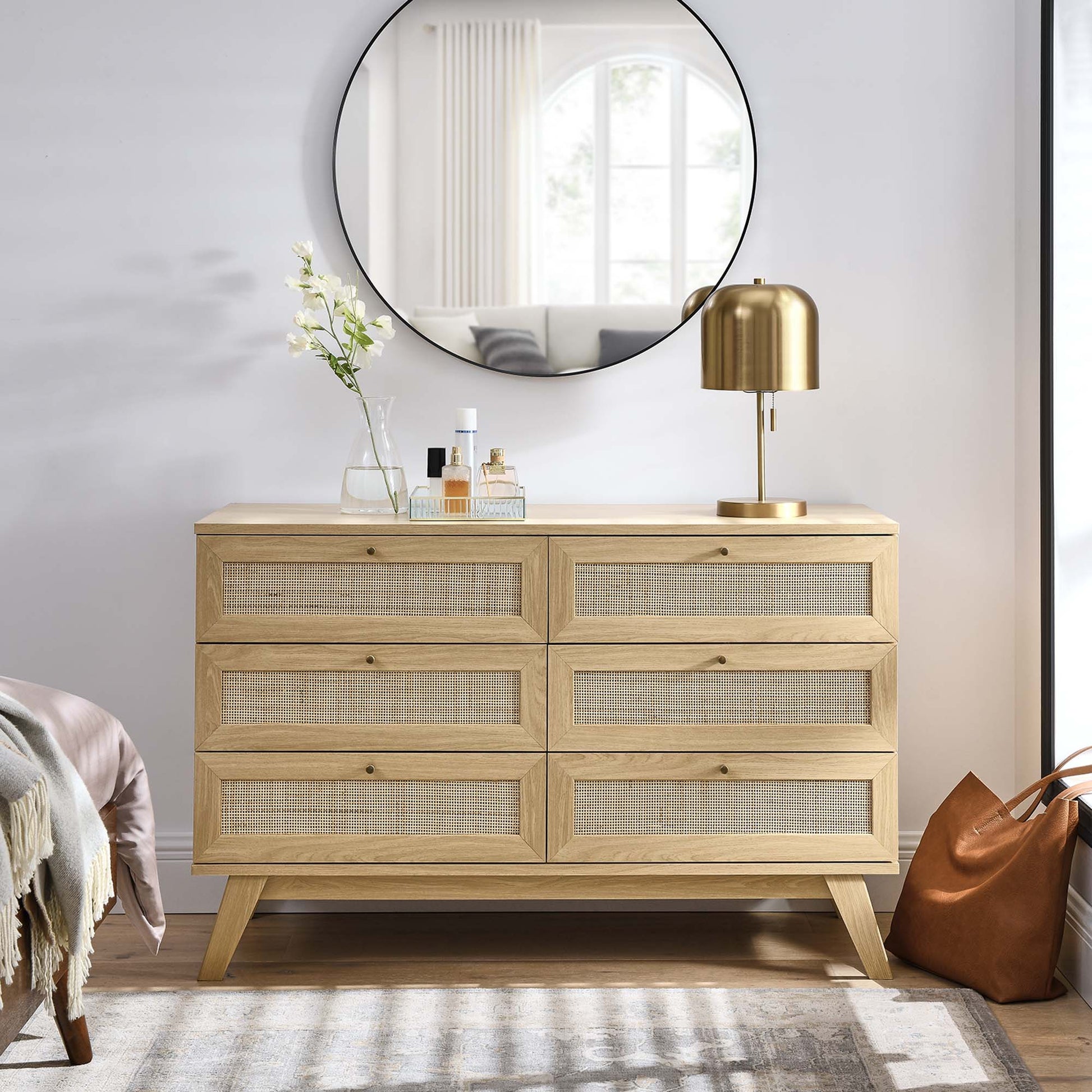 Oak dresser with six cane-front drawers & mid-century legs; brass lamp, vase & bottles on top under round mirror.