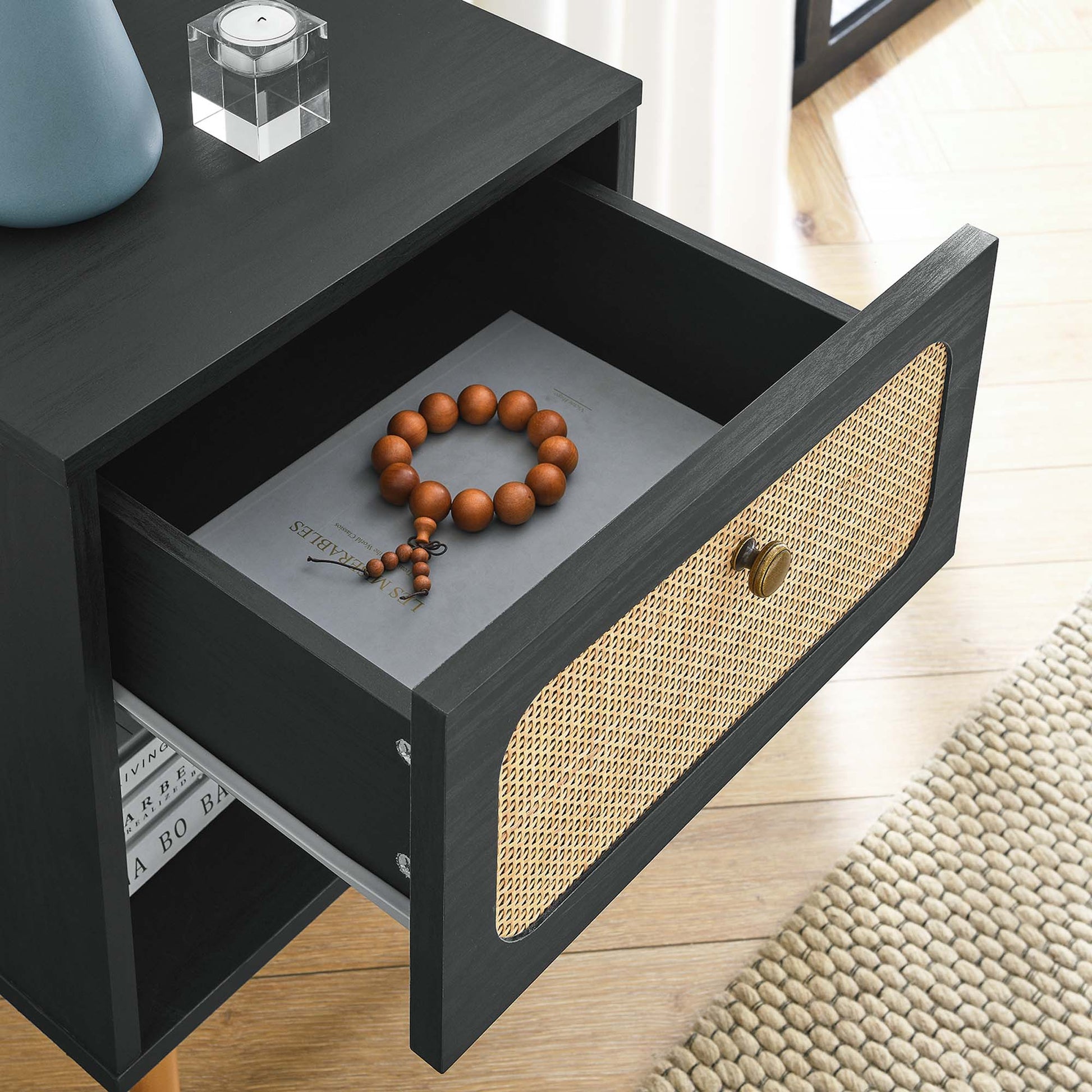 Black wooden nightstand with a woven cane front; open drawer shows a brown bead bracelet beside a book; modern rustic style.