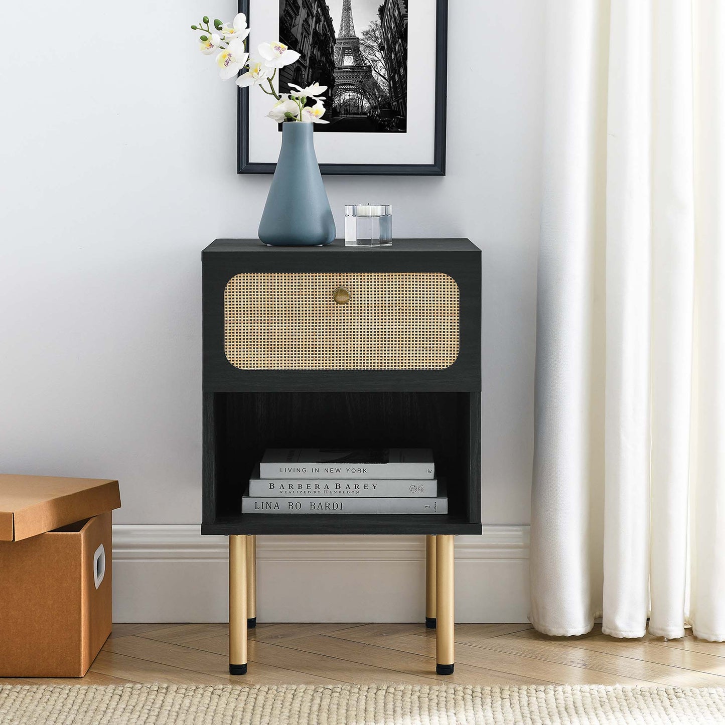 Black mid-century bedside table with woven cane drawer, open lower shelf, gold legs, topped by blue vase and white flowers.