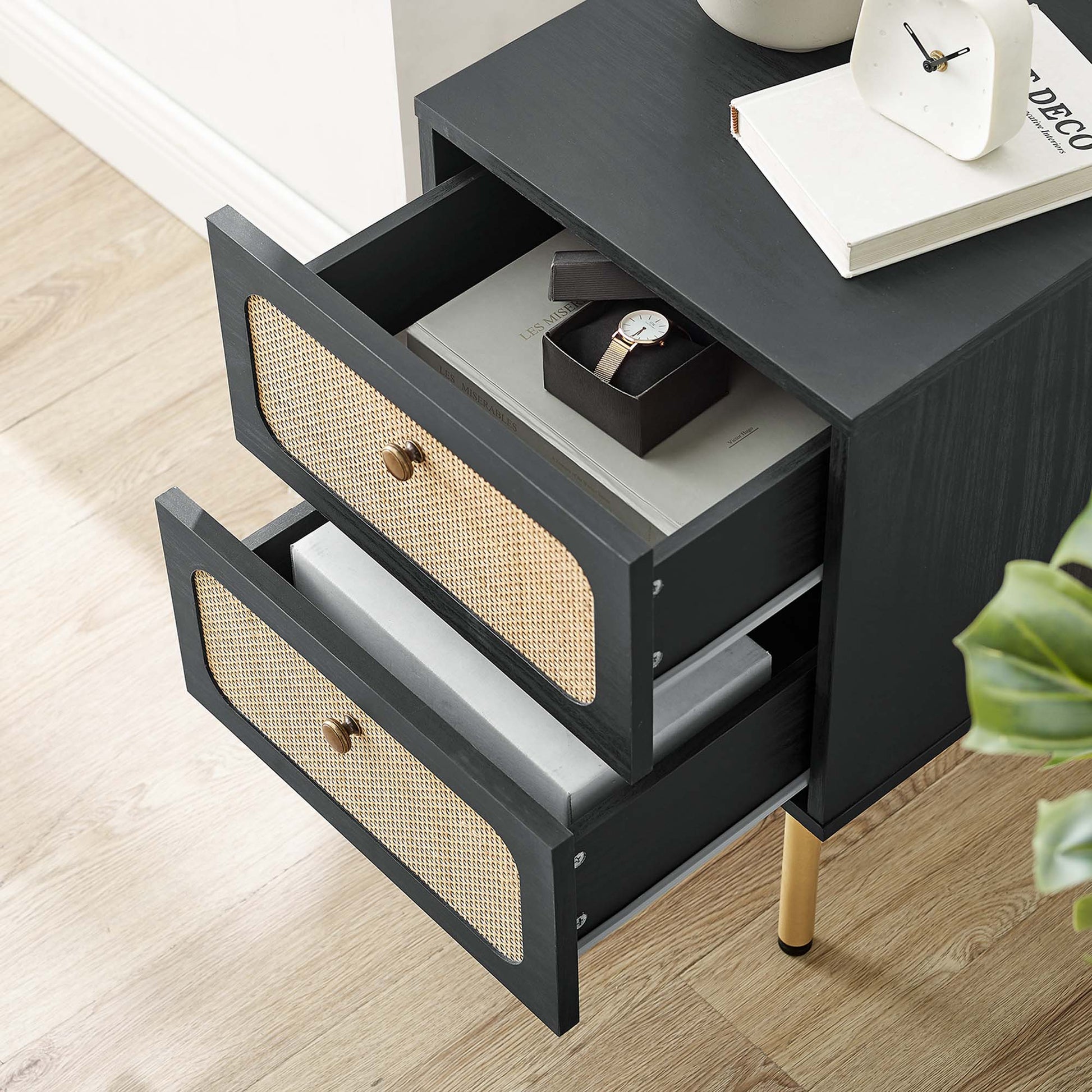 Black wood nightstand with two woven cane drawer fronts, brass knobs and gold legs; top with books and a clock.