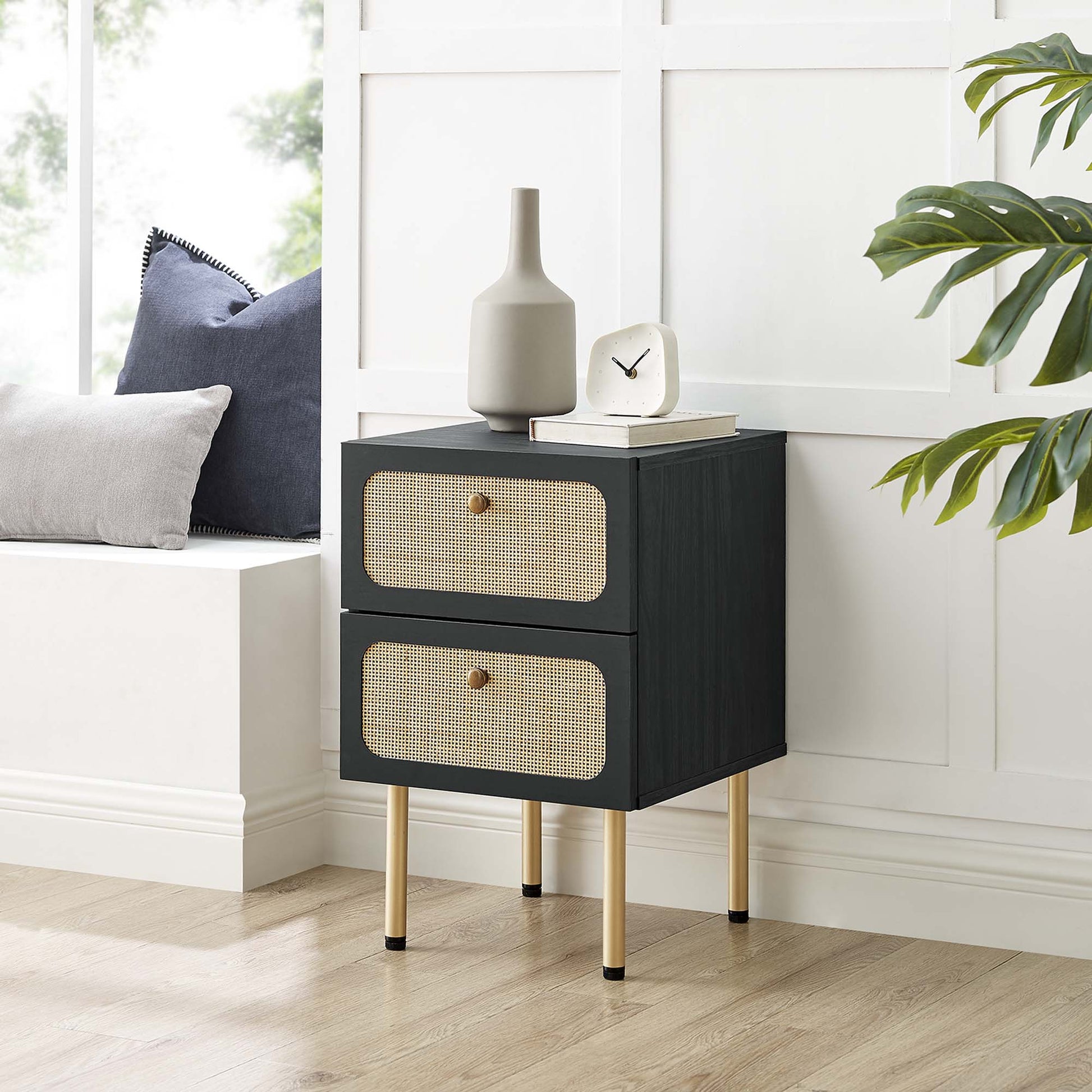 Black two-drawer bedside table with woven rattan fronts & gold legs; vase, clock & books on top.