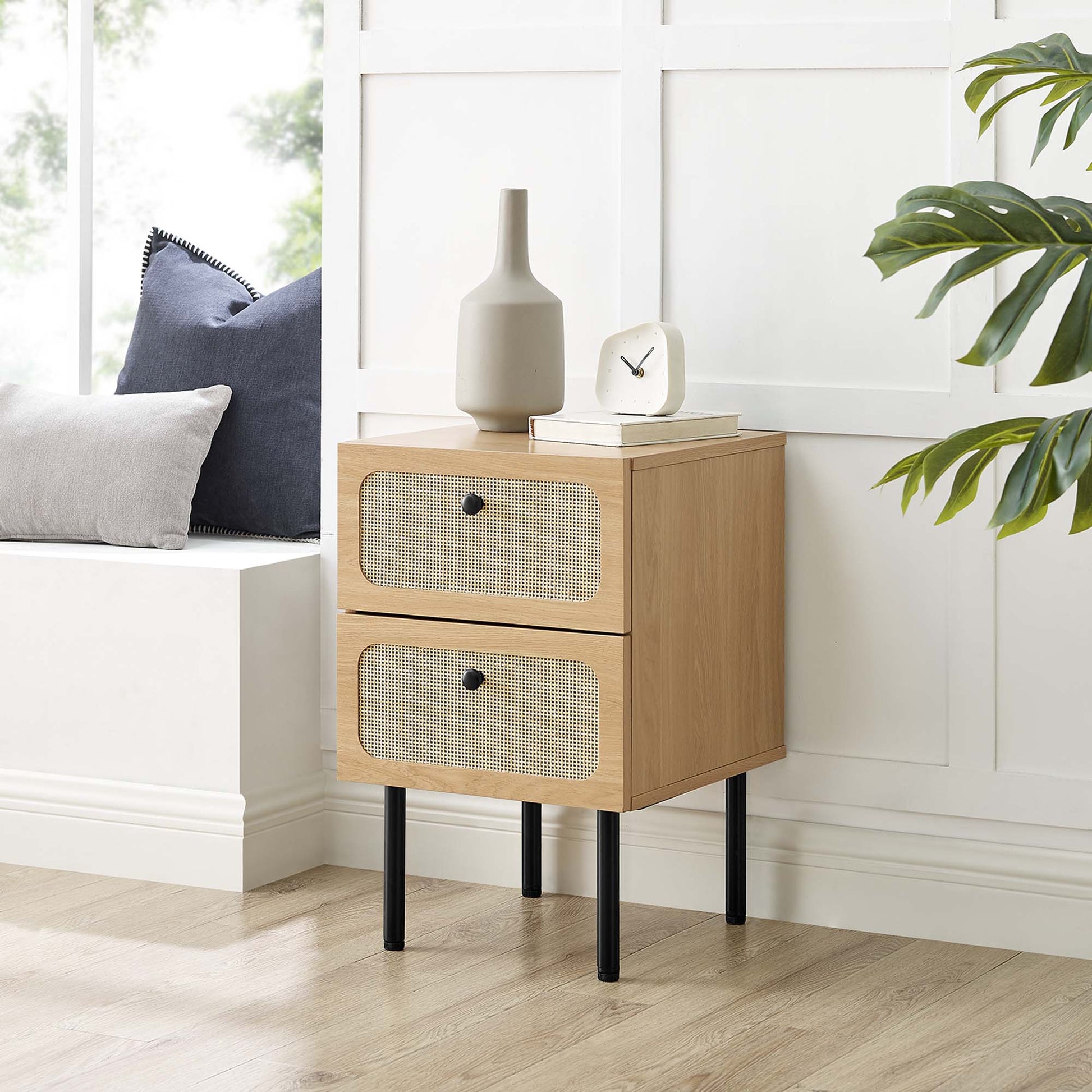 Oak wood two-drawer nightstand with woven rattan fronts on black tapered legs, decor vase, clock, books