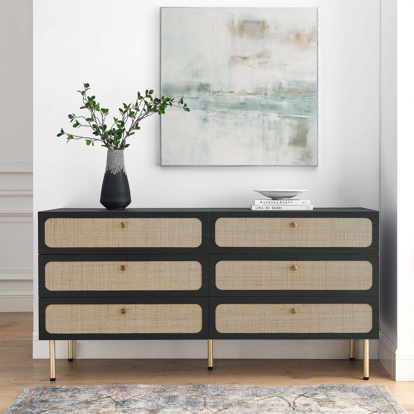 Charcoal 6-drawer dresser with cane-front drawers, gold legs and knobs, topped with a vase of greenery and books, abstract art above.