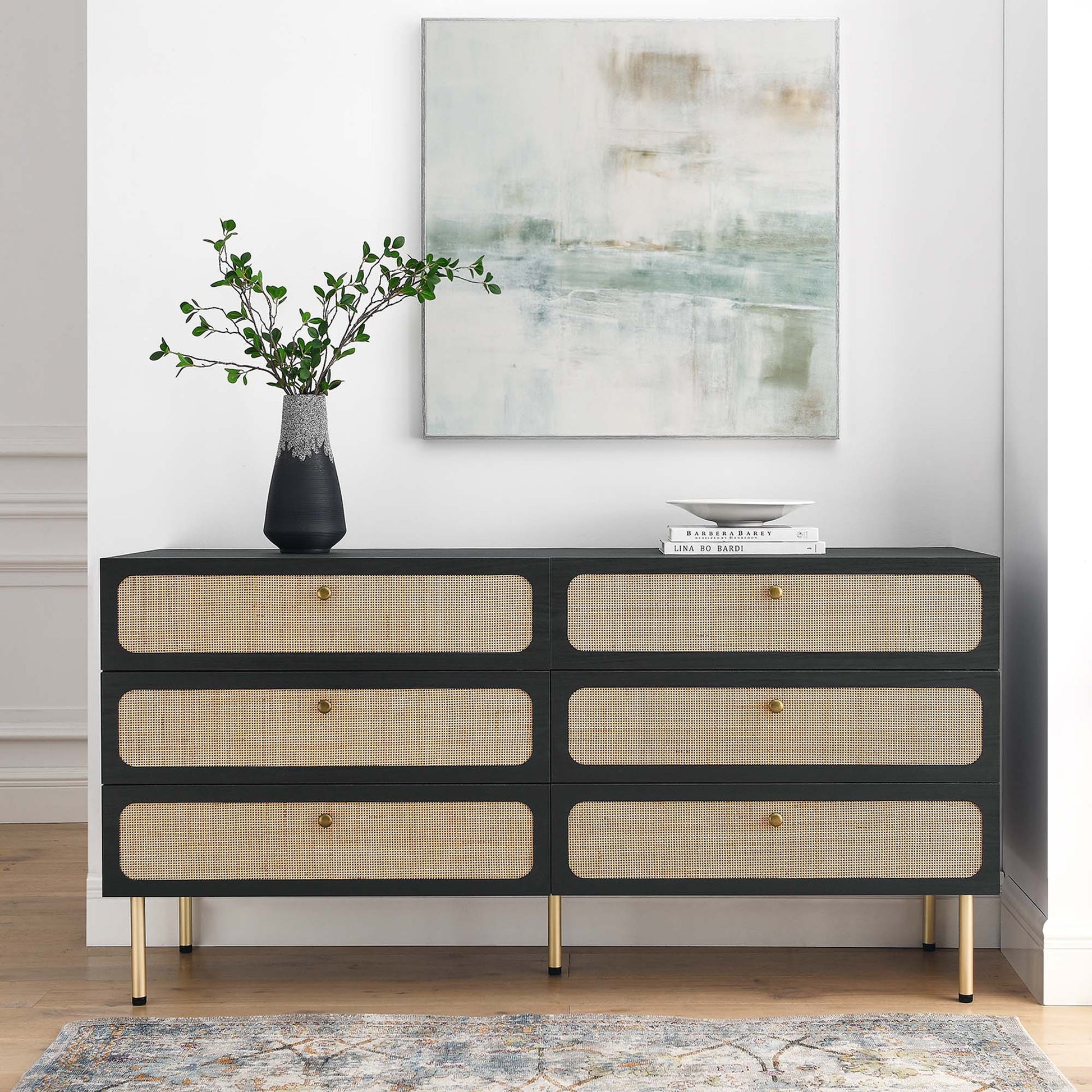 Charcoal 6-drawer dresser with cane-front drawers, gold legs and knobs, topped with a vase of greenery and books, abstract art above.