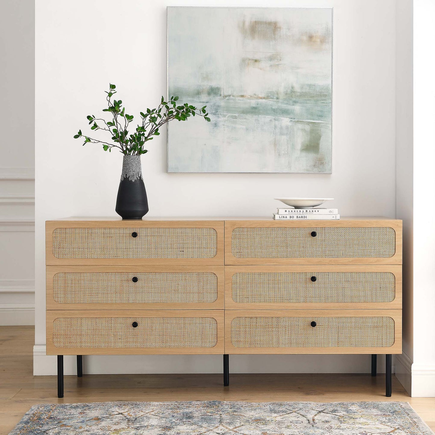 Oak 6-drawer dresser with woven-rattan fronts, black knobs and slim black legs, greenery vase, books, and abstract art.