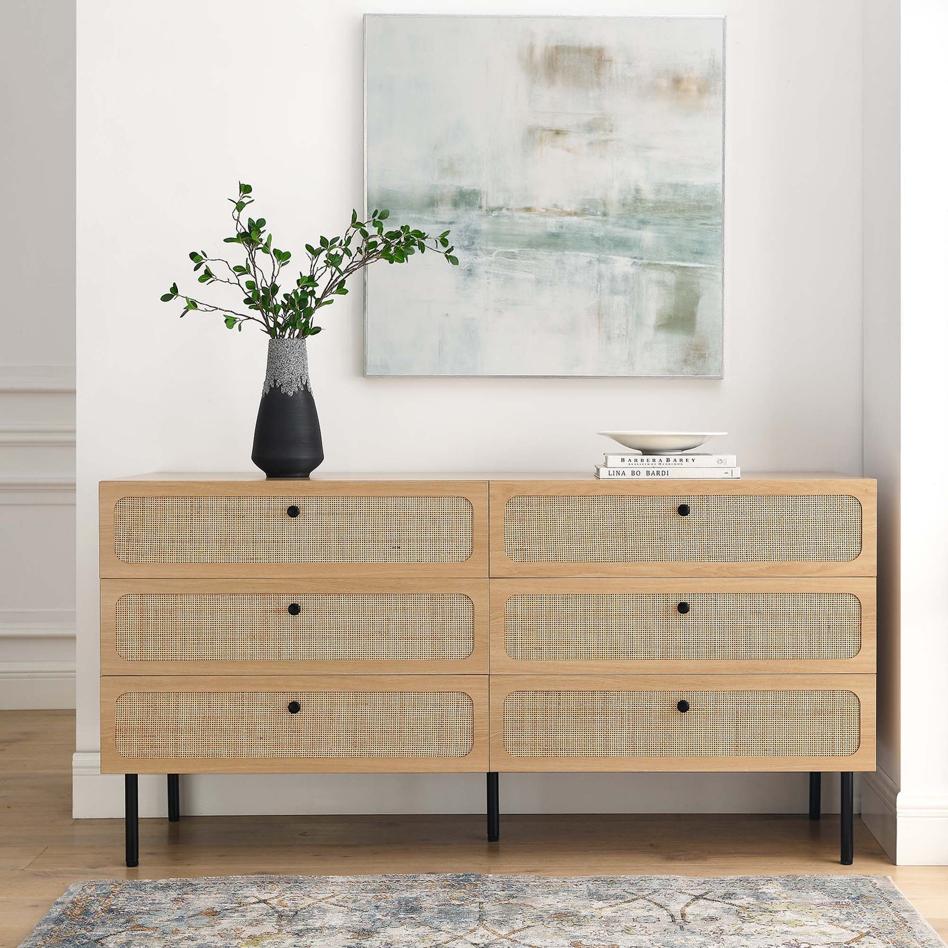 Oak 6-drawer dresser with woven-rattan fronts, black knobs and slim black legs, greenery vase, books, and abstract art.