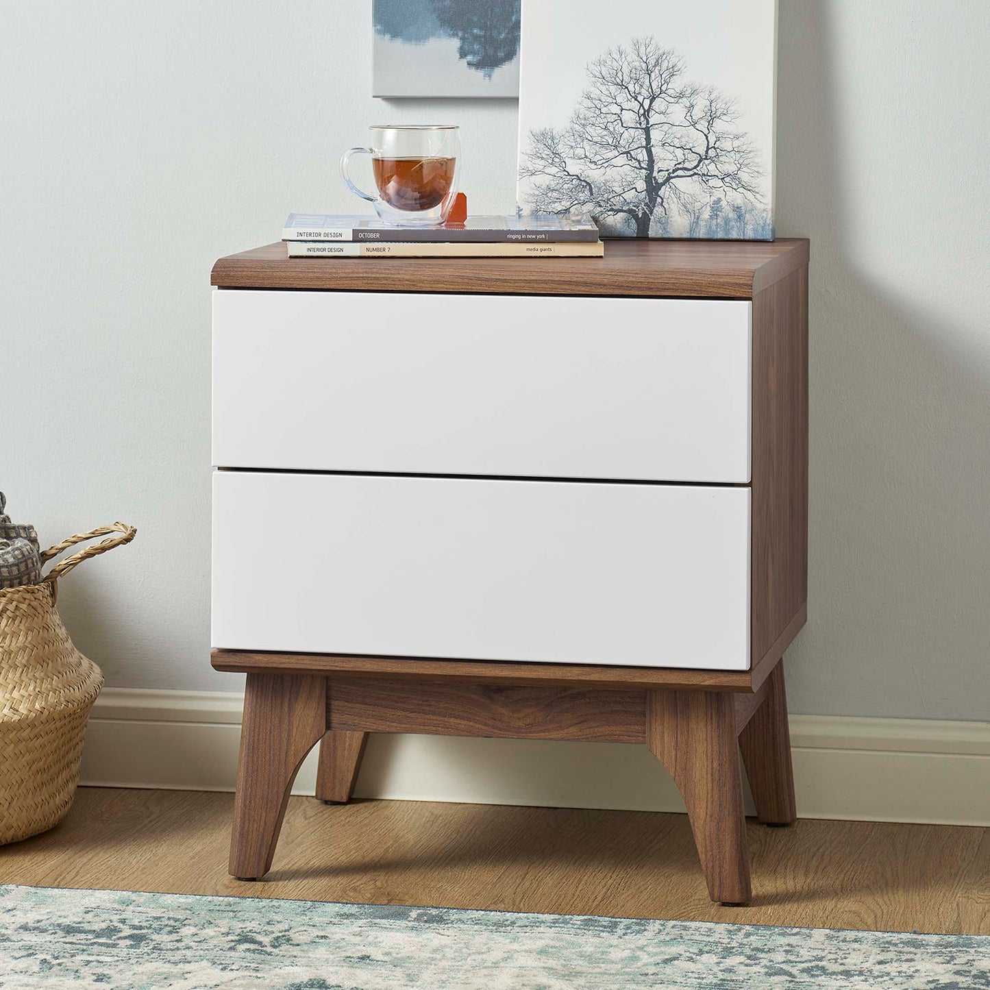 Walnut wood two-drawer nightstand with white drawer fronts, a glass teacup, books, and a tree artwork.