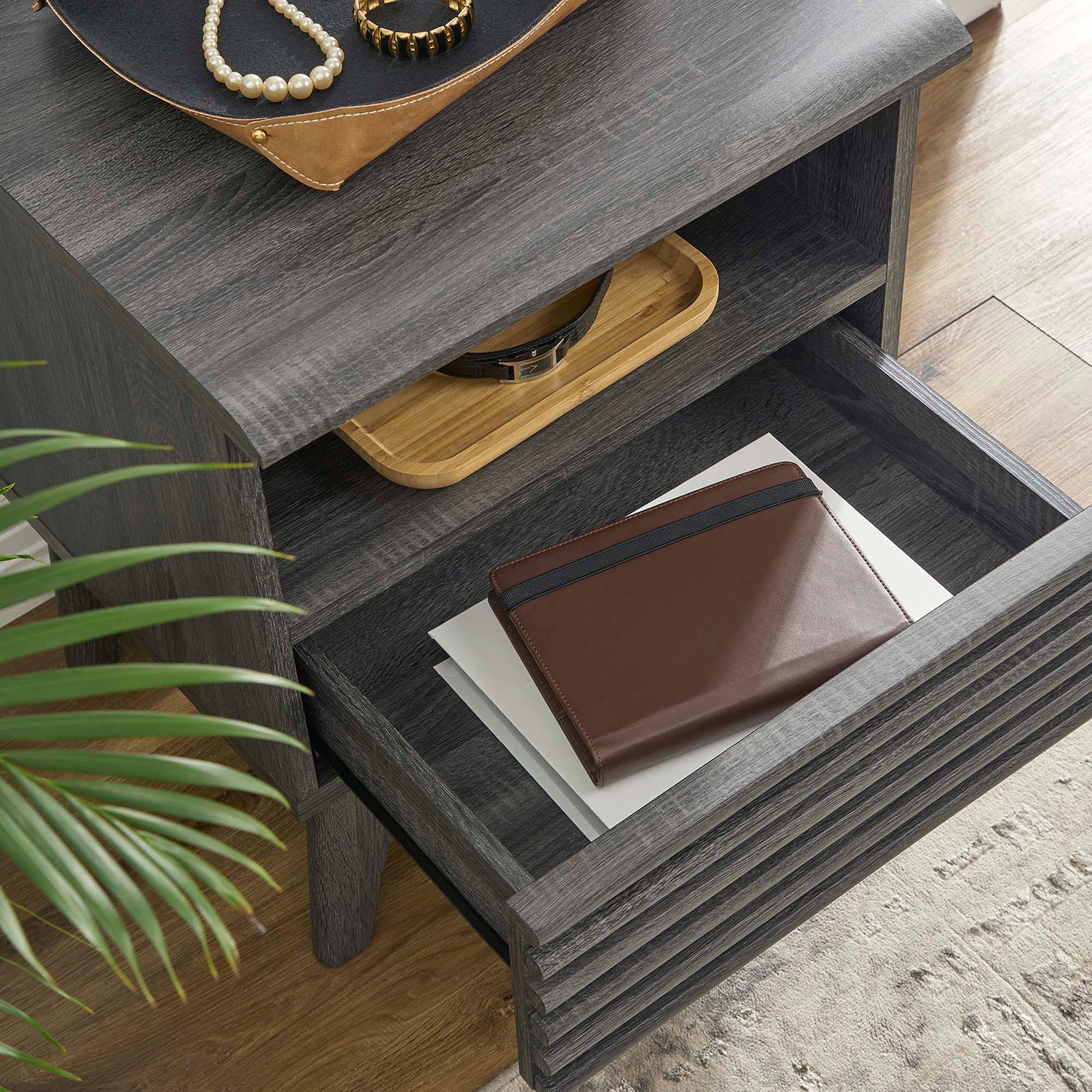 Dark gray nightstand with drawers; brown wallet on papers on top, with a wooden tray and jewelry, and a plant nearby.