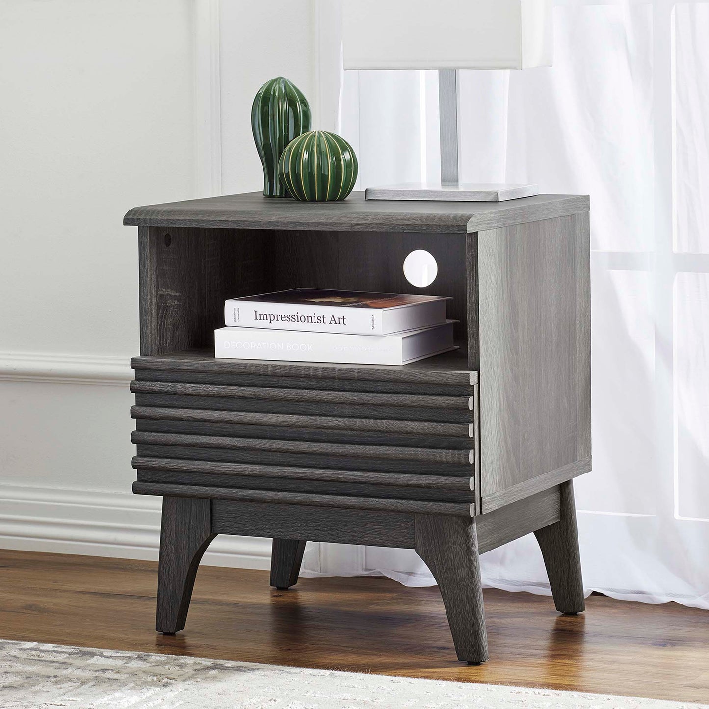 Black nightstand with a slatted drawer front, open shelf, and tapered legs; green ceramic vases and a lamp on top.